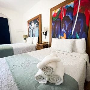 Double Double Standard room at At Wind Chimes Boutique Hotel in San Juan, Puerto Rico — two double beds with bold blue and red original artwork headwalls, sage green coverlets, and hotel-monogrammed linens
