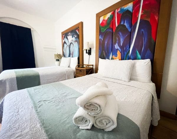 Double Double Standard room at At Wind Chimes Boutique Hotel in San Juan, Puerto Rico — two double beds with bold blue and red original artwork headwalls, sage green coverlets, and hotel-monogrammed linens