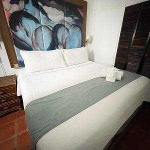 Second bedroom of the 2-Bedroom Suite at At Wind Chimes Boutique Hotel in San Juan, Puerto Rico — queen bed with original artwork headwall, sage green coverlet, and terracotta tile floors