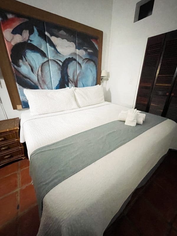Second bedroom of the 2-Bedroom Suite at At Wind Chimes Boutique Hotel in San Juan, Puerto Rico — queen bed with original artwork headwall, sage green coverlet, and terracotta tile floors