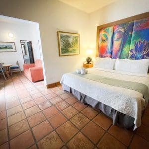 Deluxe King room view at At Wind Chimes Boutique Hotel in San Juan, Puerto Rico — colorful Buddha artwork headwall, terracotta tile floors, king bed, and separate sitting area with coral armchair