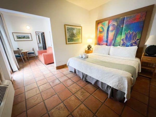 Deluxe King room view at At Wind Chimes Boutique Hotel in San Juan, Puerto Rico — colorful Buddha artwork headwall, terracotta tile floors, king bed, and separate sitting area with coral armchair