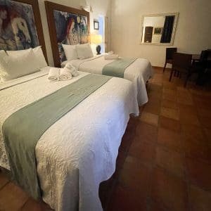 Double Double Deluxe room at At Wind Chimes Boutique Hotel in San Juan, Puerto Rico — two queen beds with original artwork headwalls, sage green coverlets, and terracotta tile floors