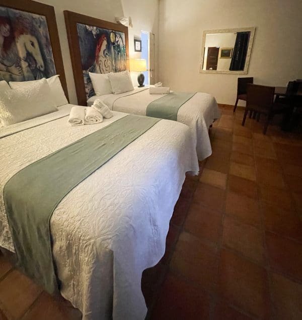 Double Double Deluxe room at At Wind Chimes Boutique Hotel in San Juan, Puerto Rico — two queen beds with original artwork headwalls, sage green coverlets, and terracotta tile floors